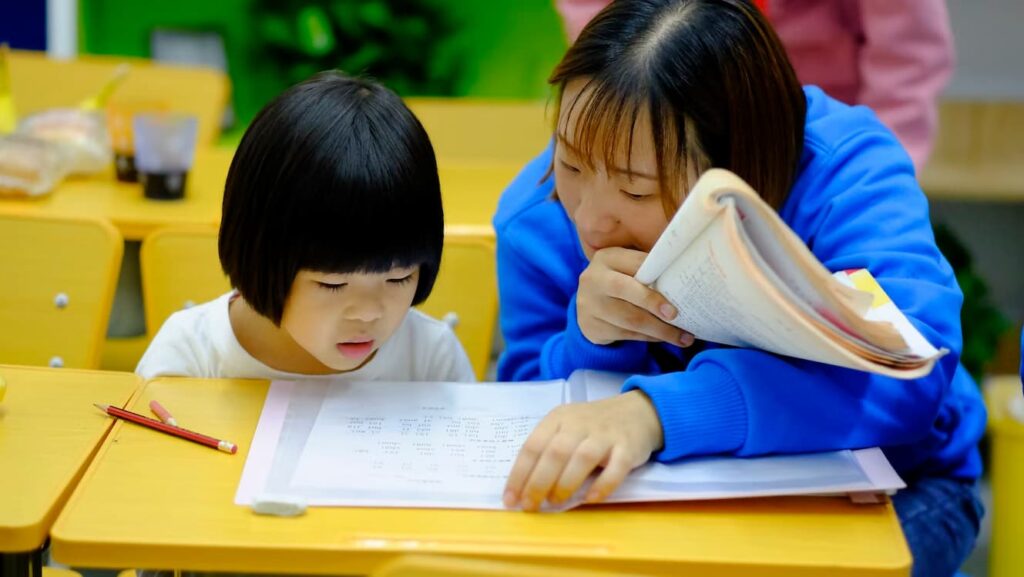 A teacher helps a young child with schoolwork at a classroom desk